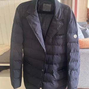 Mörkblå dunjacka kavaj från Snoot - Snygg mörkblå dunjacka i kavajmodell från Snoot. Jackan har klassisk krage, tre framfickor och diskret Snoot-logga på ärmen. Perfekt för dig som vill ha en stilren look med extra värme. Fodrad och quiltad för maximal komfort. Ny pris 4000