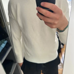Vit stickad tröja med rund hals - Säljer en stilren vit stickad tröja med rund hals och långa ärmar. Tröjan har ribbade muddar vid ärmslut och nederkant, vilket ger en snygg och enkel look. Perfekt för dig som gillar minimalistisk stil och vill ha något mjukt och bekvämt. Jag är 181cm och 75kg