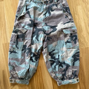 Replay cargopants i camomönster - Säljer ett par cargopants från Replay med brunt, grönt och svart camomönster. Byxorna har flera stora fickor med lock och knappar och knäppning vid bensluten. Perfekta för dig som gillar streetwear. Rökfritt hem. Storlek w27 i midjan.