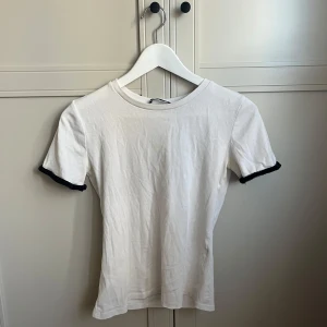 Vit topp med svarta virkade ärmslut - Säljer en vit topp med korta ärmar och svarta virkade detaljer längst ut på ärmsluten. T-shirten har en klassisk rund halsringning. Perfekt för dig som gillar enkla plagg med en twist! Det är även en av sömmarna som är sprucken men syns inte från utsidan