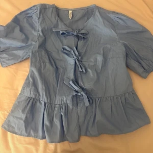 Blå blus med knyt och volang - Supersöt ljusblå blus med tre knyt framtill och volang nertill. Puffiga korta ärmar och rund halsringning ger en romantisk vibe. Perfekt att styla med jeans eller kjol för en chill look.