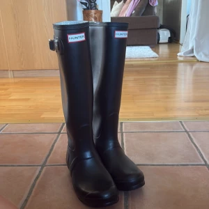 Svarta Hunter stövlar - Säljer ett par klassiska svarta Hunter stövlar med högt skaft och rund tå. Stövlarna har en snygg spänndetalj på sidan och är tillverkade i slitstark syntet som tål regn och lera. Perfekta för dig som vill ha en stilren look även när det är blött ute.