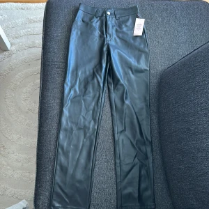 Svarta raka byxor i skinnimitation med etikett kvar  - Snygga svarta byxor i skinnimitation med raka ben och klassisk femficksdesign från Nelly. Byxorna har hällor för bälte och stängs med knapp och dragkedja. 