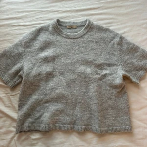 Grå stickad tröja Gina Tricot - Mjuk och chill grå stickad tröja från Gina Tricot med korta ärmar och rund hals. Tröjan har ribbade detaljer vid hals, ärmslut och nederkant. Perfekt för lager-på-lager och funkar till både jeans och kjol. Enkel och clean look som är lätt att matcha.