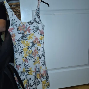 Blommig kortklänning från H&M Divided - Supersöt kortklänning från H&M Divided med blommigt mönster i gult, rosa och svart på vit botten. Klänningen är ärmlös med rundad hals och har en lätt utställd kjol. Perfekt för varma dagar och ger en fräsch och ungdomlig vibe.