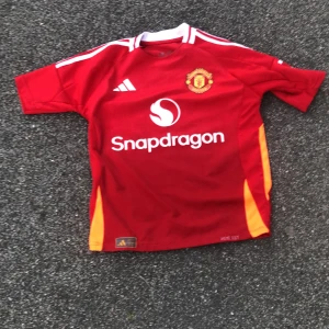 Manchester United Garnacho #17 tröja - Säljer en officiell Manchester United fotbollströja från Adidas med Garnacho och nummer 17 på ryggen. Tröjan är röd med vita detaljer och orange inslag på sidorna, har korta ärmar och klubbmärke på bröstet. Materialet är lätt och andas, perfekt för match eller träning.