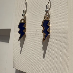 Blå blixtformade örhängen - Snygga örhängen i form av blixtar med mörkblå insida och guldfärgad kant. Perfekt för dig som vill sticka ut med en cool och unik accessoar. Hängande modell med metallkrok, passar till alla tillfällen när du vill addera lite edge till din stil.