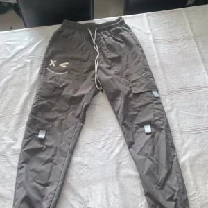 Grå cargopants med snörning - Säljer ett par grå cargopants med coola detaljer och tryck på ena benet. Byxorna har flera fickor, snörning i midjan och reflekterande band på benen. Perfekta för dig som gillar streetstyle och vill ha något bekvämt och snyggt.