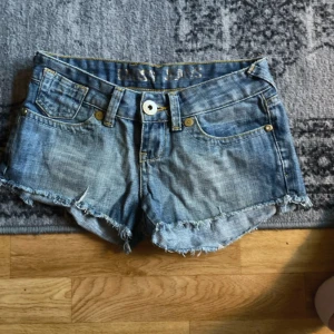 Blå jeansshorts från Cross Jeans - Snygga blå jeansshorts från Cross Jeans med låg midja och råa, fransiga kanter. Klassisk femficksmodell med guldfärgade detaljer och broderade hjärtan på bakfickorna. Storlek 25. Skriv vid prisförslag 