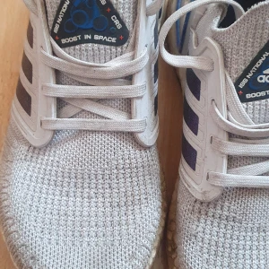 Adidas Ultraboost ISS National Lab sneakers - Adidas Ultraboost ISS National Lab sneakers i ljusgrå stickad mesh med blå metallic ränder på sidorna och vit boost-sula. Skorna har snörning och svart Continental-yttersula för extra grepp. Perfekt för dig som gillar coola detaljer och bekväm stil.