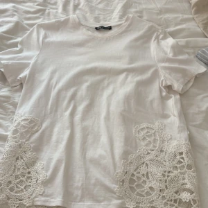 Vit topp med spetsdetaljer från Zara💕 - Vit topp från Zara i bomull med korta ärmar och rund hals. Nedtill finns snygga virkade spetsdetaljer på båda sidor som ger en unik och romantisk touch. Perfekt för dig som gillar stilrena plagg med det lilla extra.❤️