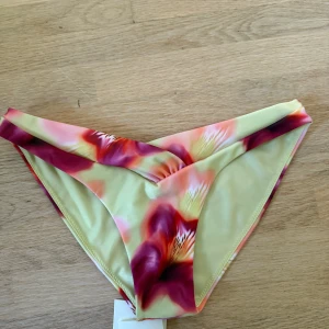 Blommig bikini från Peppermayo - Superfin bikini från Peppermayo med blommigt mönster i rosa, lila och gula toner. Bikinin har en twistad bandeau-topp och matchande högt skurna bikinitrosor med omlott-detalj. Perfekt för stranden och sommarens alla poolhäng!