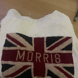   Morris tröja med flagga - Snygg stickad tröja från Morris i storlek Large. Tröjan är vit med ett stort Union Jack-flagga mönster i rött, svart och beige på bröstet, där det även står 'Morris'. Rund hals och långa ärmar, perfekt för dig som gillar klassisk stil med en twist.