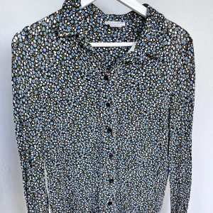 Blommig skjorta från Cooperative  - Snygg skjorta från Cooperative med småblommigt mönster i blått, vitt och gult på svart botten. Skjortan har långa ärmar, klassisk krage och knäppning framtill. Perfekt för dig som gillar färg och mönster!