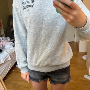 Grå hoodie med tryck och huva - Säljer en ljusgrå hoodie med svart texttryck 'JE M'EN AMOUR' och en stjärna både fram och bak. Tröjan har huva, ribbade muddar och är i mjukt bomullsmaterial. Perfekt för chill dagar och enkel att matcha med jeans eller shorts.