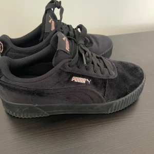 Svarta Puma sneakers i mocka, strl 36 - Säljer ett par svarta Puma sneakers med SoftFoam+ innersula för extra komfort. Skorna har snygga detaljer i rosa på sidan och plösen, klassisk snörning och en platt sula. Ovandelen är i mjuk mocka som ger en schysst look och känsla.