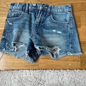 Blå jeansshorts med fransar från Zara - Snygga blå jeansshorts från Zara med råa, fransiga kanter och slitna detaljer. Klassisk femficksmodell med knapp och dragkedja framtill. Perfekt för dig som gillar en avslappnad och cool stil på sommaren.