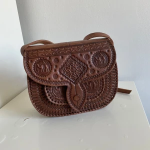 Brun axelväska i skinn med mönster - Säljer en brun axelväska i skinn med unika, handgjorda mönster och detaljerade präglingar på locket. Väskan har en klassisk form, lång axelrem och snygga sömmar runt kanterna. Perfekt för dig som vill ha något som sticker ut och känns genuint.