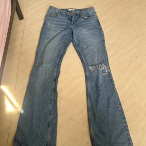 Blå bootcut jeans från Gina tricot - Bootcut jeans i klassisk blå denim från Perfect Jeans, storlek 38. Byxorna har slitna detaljer och hål på ena benet för en edgy look. Tillverkade i bomull med femficksmodell och normal passform. Perfekta för dig som gillar en avslappnad och trendig stil.