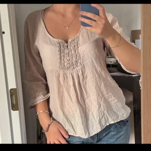 Beige blus med knappar från NY Garderob - Säljer en ljusbeige blus från NY Garderob med snygga knappar och broderade detaljer framtill. Blusen har en rundad halsringning och trekvartsärmar med knyt vid ärmslutet. Tillverkad i mjukt linne, perfekt för vår och sommar.