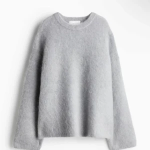 Ljusgrå stickad tröja från H&M - Mjuk och fluffig ljusgrå stickad tröja från H&M med rund hals och långärmad passform. Tröjan har en loose fit och är perfekt att bära över en skjorta. Materialet är en skön blandning med ull som ger en mysig känsla.