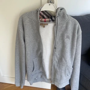 Burberry hoodie med rutig huva - Två hoodies från Burberry, en grå och en marinblå, båda med dragkedja och klassiskt rutigt foder i huvan. Mjukt bomullsmaterial, fickor fram och broderad logga på bröstet. 1500kr st eller 2500kr för båda