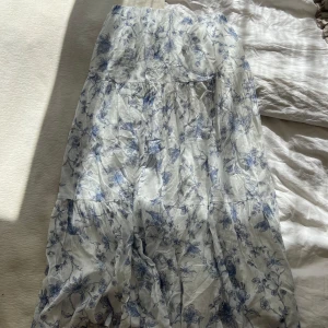 Blommig långkjol från Gina Tricot - Luftig långkjol från Gina Tricot i vitt med blått blommigt mönster. Kjolen har resår i midjan och är tillverkad i mjuk bomull, perfekt för varma dagar. Den har en flowy passform och fina volangdetaljer som ger extra rörelse. Tyvärr så har den gått lite sönder som ni kan se på bilden men det är inget som inte går att laga, säljer då den är för liten men super fin kjol! Pris kan diskuteras vid snabb affär💞