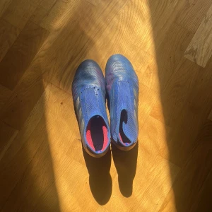 Blå adidas fotbollsskor utan snörning - Säljer ett par blå fotbollsskor från adidas med silvriga ränder på sidorna och röda detaljer. Skorna har en strumpliknande passform utan snörning och är tillverkade i syntetmaterial. Pris kan såklart diskuteras vid snabb affär!