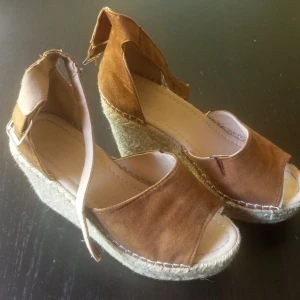 Bruna sandaler med platåsula - Snygga sandaler i brun mocka med öppen tå och hög platåsula i repimitation. Rem runt vristen med spänne för extra support. Perfekta för sommarens alla outfits och ger skön höjd utan att tumma på komforten.