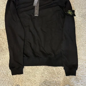 Svart Stone Island sweatshirt - Svart sweatshirt från Stone Island med klassisk rund hals och ribbade muddar. Tröjan har den ikoniska Stone Island-loggan på vänster ärm och är tillverkad i mjuk bomull. Perfekt för en clean och stilren look.