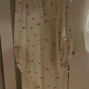 Vit långklänning med röda blommor från Adoore - Säljer en superfin vit långklänning från Adoore med små röda blommor. Klänningen har långa puffärmar med resår vid handlederna och knappar framtill. Perfekt för dig som gillar romantisk stil och vill sticka ut med ett gulligt mönster.