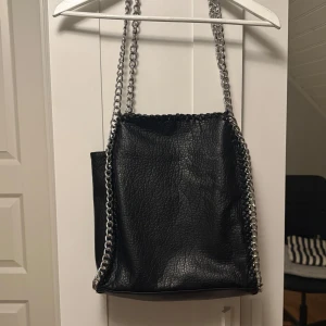 Svart axelväska med kedjedetaljer - Snygg svart axelväska i skinnimitation med grova silverfärgade kedjor som axelrem och längs kanterna. Väskan har en cool, lite boxig form och passar perfekt för dig som gillar en edgy stil.