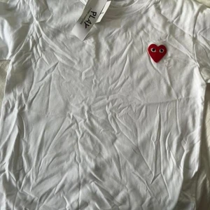 Vit t-shirt Comme des Garçons Play strl 150 - Vit t-shirt från Comme des Garçons Play med det ikoniska röda hjärtat broderat på bröstet. Klassisk rund hals och korta ärmar. Tillverkad i mjuk bomull som känns skön mot huden. Perfekt för dig som gillar stilrena plagg med en twist.