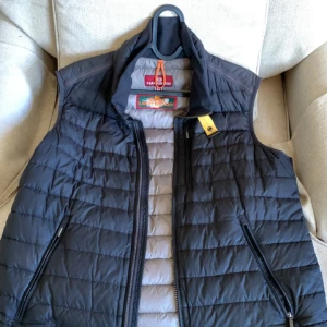 Svart dunväst från Parajumpers - Säljer en svart dunväst från Parajumpers med quiltad design och två dragkedjefickor. Västen har hög krage, snygga detaljer med logga och orange ögla vid nacken. Lättviktig och perfekt att slänga över en hoodie eller tröja.