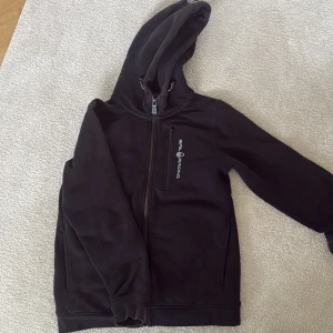Svart fleecejacka Sail Racing 150 - Svart fleecejacka från Sail Racing med huva och hel dragkedja. Jackan har en bröstficka med dragkedja och logga på bröstet. Mjuk insida och två sidofickor, perfekt för kyliga dagar. Enkel och stilren design som funkar till det mesta.