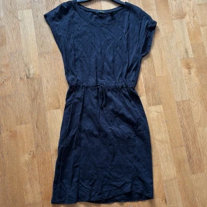 Svart vardagsklänning med snörning - En svart, enkel och stilren kortklänning från Vero Moda med rund hals och korta ärmar. Klänningen har snörning i midjan för en snygg passform och är tillverkad i mjuk bomull. Perfekt för dig som gillar minimalistisk stil och vill ha något bekvämt och lättburet. Väldigt snygg vardagsklänning.