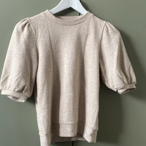 Beige puffärm tröja - Säljer en beige tröja med puffiga korta ärmar och rund hals. Materialet är mjukt och har en lite fluffig struktur som ger en mysig känsla. Perfekt för dig som gillar enkel men ändå trendig stil. Passar dig som vill ha något chill men ändå snyggt. Änvänd fåtal gånger. Köptes för ca. 150kr. pris kan förhandlas.