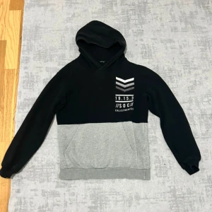 Svart och grå hoodie från SOC - Snygg hoodie från SOC i svart och grått med tryck på bröstet. Tröjan har huva, känguruficka och långa ärmar. Perfekt för chill dagar eller när du vill ha en sportig look. Mjuk insida och klassisk passform.