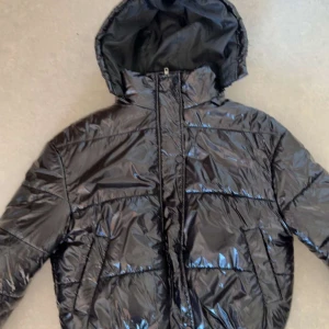 Svart pufferjacka från Boohoo - Snygg svart pufferjacka från Boohoo med glansig finish och huva. Jackan har dragkedja framtill, quiltade detaljer och lång ärm. Perfekt för kalla dagar när du vill ha en cool och trendig look.