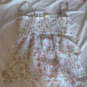 Blommig volangtop med smock och axelband - Supersöt blommig linnetopp med volanger och smock upptill. Tunt, luftigt tyg i vitt med rosa och gröna blommor. Smala axelband och flowy passform, perfekt för varma dagar. Ger en romantisk och somrig vibe.