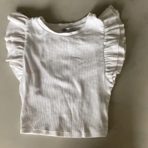 Vit volangtopp från Zara - Supersöt vit topp från Zara med volangdetaljer på axlarna. Toppen är ribbad och har en rak, croppad passform. Perfekt för dig som gillar en feminin och trendig stil. Tillverkad i mjuk bomull som känns skön mot huden.