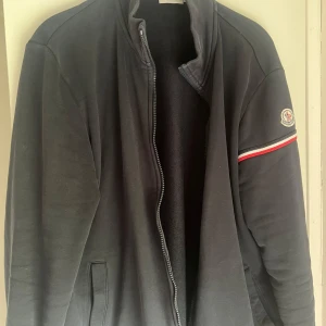 Marinblå Moncler jacka med dragkedja - Marinblå jacka från Moncler med hög krage och hel dragkedja framtill. Klassisk Moncler-logga på ärmen och en rödvit randdetalj. Jackan har två fickor och är tillverkad i mjukt bomullsmaterial. Perfekt för dig som gillar stilren och sportig look. Använd men är i ett mycket bra skick! Pris tänker jag runt 1200-1300 men kan förhandlas! Storleken är XL men är som en L.