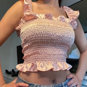 Rosa smockad satin tubtopp med volang - Supersöt ljusrosa tubtopp i glansig satin med smockad midja och volangdetaljer upptill och nedtill. Axelbanden är avtagbara och i samma material, så du kan bära toppen både med och utan axelband. Perfekta festtoppen❤️