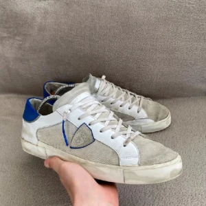 Beige och vita sneakers med blå detalj - Snygga sneakers från Philippe Model i beige mocka och canvas med vita läderdetaljer och blå häl. Klassisk låg modell med snörning och rund tå. Perfekta för en avslappnad och trendig stil.