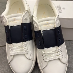 Valentino Garavani Open sneakers vit/marin - Snygga Valentino Garavani Open sneakers i vitt skinn med bred marinblå läderdetalj över mitten. Klassisk rund tå, vita snören och ikoniska nitar baktill på sulan. Perfekta för dig som gillar stilrena och lyxiga sneakers med en twist.