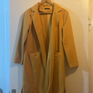 Gul lång kappa  - Snygg och stilren gul kappa med klassisk krage och två stora fickor framtill. Lång modell som ger en cool och trendig look, perfekt för att sticka ut i mängden. Enkel att matcha med olika outfits.