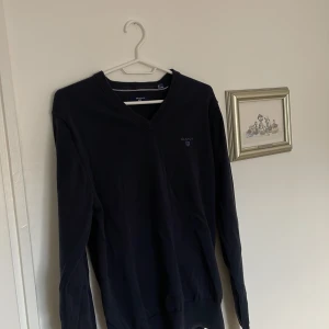 Svart tröja från GANT - Säljer en stilren svart tröja från GANT i premium bomull. Tröjan har en v-ringad design och långa ärmar, perfekt för en klassisk look. GANT-loggan är diskret broderad på bröstet. Passar bra till både jeans och chinos.