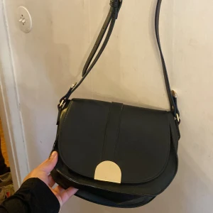 Svart axelväska med gulddetalj - Snygg svart axelväska i skinn med justerbar axelrem och elegant gulddetalj på framsidan. Väskan har en praktisk dragkedja inuti för säker förvaring. Perfekt för att ge en stilren touch till din outfit.