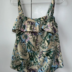 Blommig linne med volangdetalj - Säljer ett somrigt linne med blommigt mönster i grönt, blått och lila på en beige bakgrund. Toppen har smala axelband och en söt volangdetalj vid bysten. Perfekt för varma dagar!