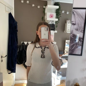 Vit ribbad topp med svarta detaljer - Säljer en stilren vit ribbad topp med svarta volangdetaljer vid kragen och ärmsluten. Toppen har trekvartsärmar och en knäppning framtill. Perfekt för en chic och avslappnad look.